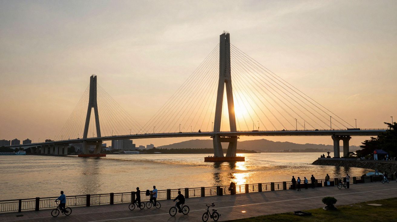 Ponte suspensa sobre o rio ao pôr do sol, com pessoas a passear de bicicleta na margem.